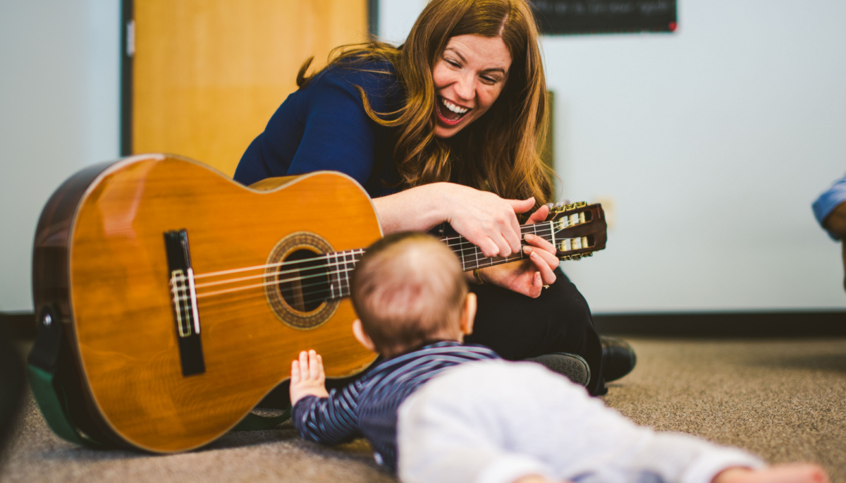 Ms. Pizzi plays the guitar as a young baby crawls on his belly and touches the guitar with his hand.