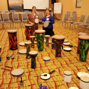 Music therapists kneel on the ground in front of a variety of hand drums, rhythm instruments, tubanos, and djembes.
