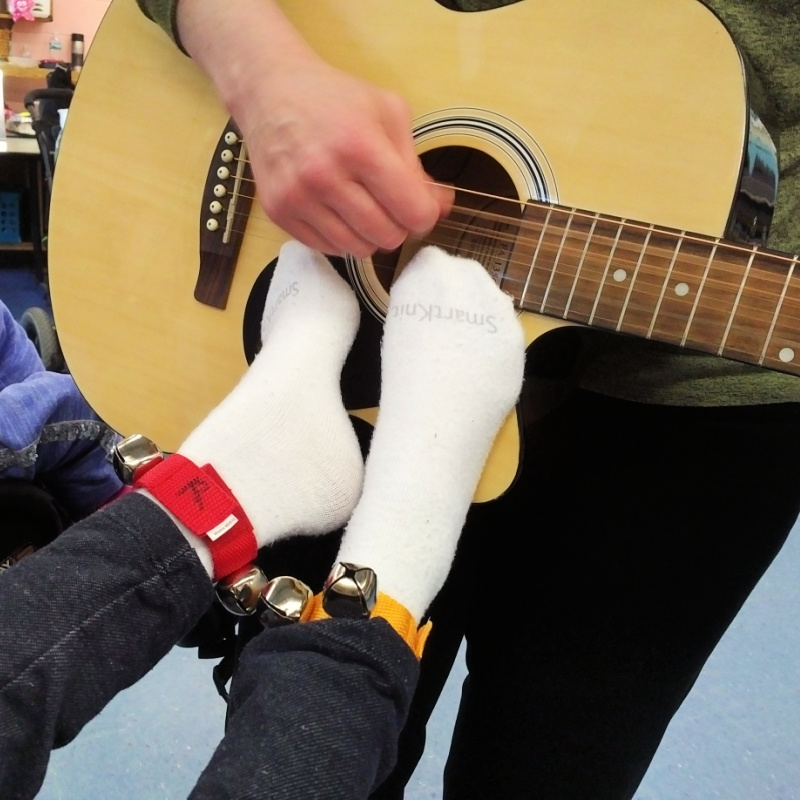 A music therapy intern holds and plays an acoustic guitar in front of a student. The student has jingle bells around his ankles and holds his feet against the body of the guitar to feel the vibrations.