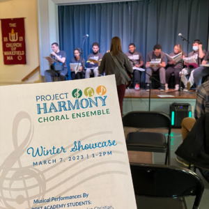 A recent Project Harmony Choral Ensemble Showcase. Ensemble members sit on a stage with open music binders in their hands as their music therapist leads them in singing, playing instruments, and soloing.